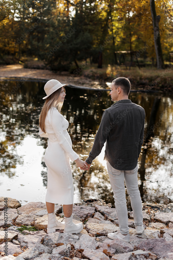 Fototapeta premium Rear view of stylish young couple in love holding hands by the pond at autumn park. Woman in knit white dress and hat and man have dating on valentine's day. Happy family love concept