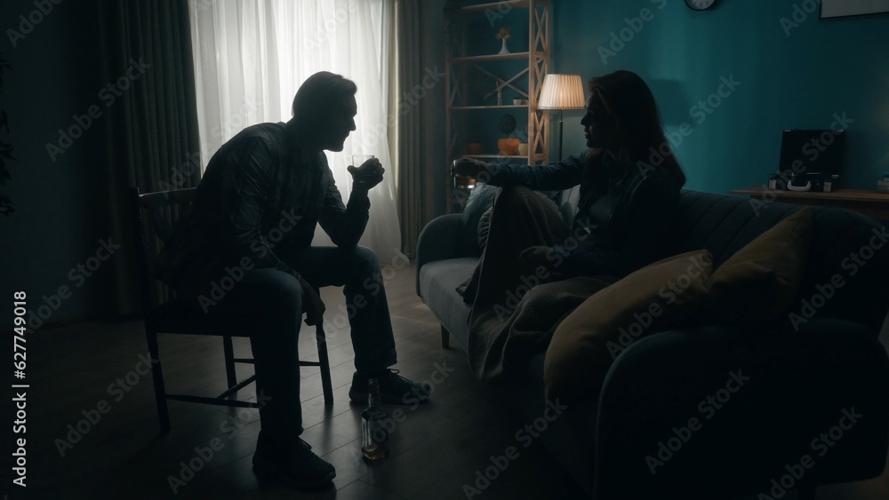 A couple sits with glasses of alcohol in a room in darkness. A man and a woman sit opposite each other, between them a bottle of alcohol. The couple escapes from problems with the help of alcohol.
