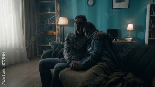 A couple is sitting on a sofa in a dark room. A man holds a woman by the hand, hugs. Support for the couple, joint problem solving, reconciliation and understanding in relationships. © kinomaster