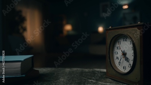 A vintage clock stands on a table against a blurred background of a room plunged into darkness close up. Time is running out and it cannot be returned, the present becomes the past.