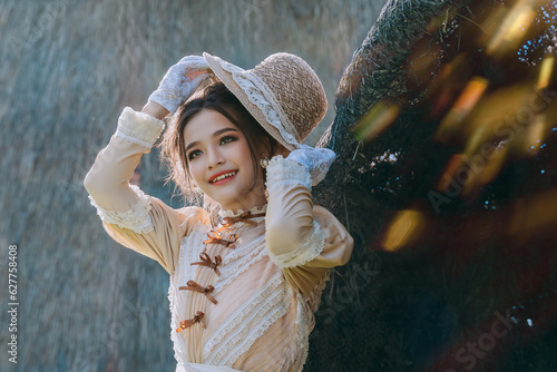 Fototapeta Young Asian woman in edwardian style dress and big hat standing in the front of the hut