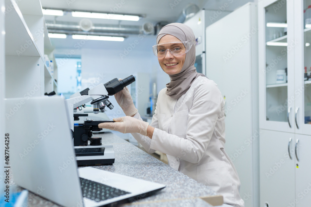 Young Muslim woman in hijab, scientist, chemist works in laboratory behind a microscope. Sitting at the table with a laptop, smilingly looking at the camera. Young Muslim woman in hijab, scientist, chemist works in laboratory behind a microscope. Sitting at the table with a laptop, smilingly looking at the camera.