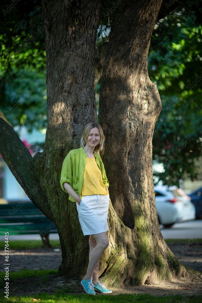 A woman in a white skirt stands near a tree in a city square.