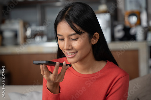 Young asian woman using voice search on smartphone in living room at home. She enjoy vocal order to help chatbot or social media.