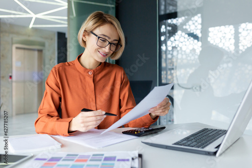 Young successful business woman working inside office with documents, woman satisfied with achievement results reviewing contract, bills and documents, financier in glasses smiling.