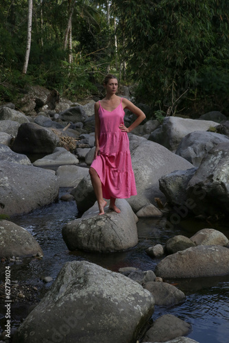 Portrait of beautiful young woman in bright pink dress posing on the large stones in the riverbed. Concept of wellbeing  mindfulness  connecting with nature and enjoying the healing sound of water