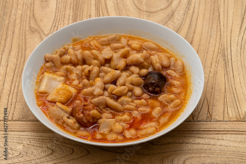 a white plate of beans with chorizo, black pudding and bacon with its broth, on a wood table