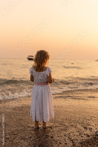 A little girl wearing a long white dress stands at the sea and watches the waves and the sunset. Sea vacation, childhood concept. A girl with long fair hair on the beach at the sunset.