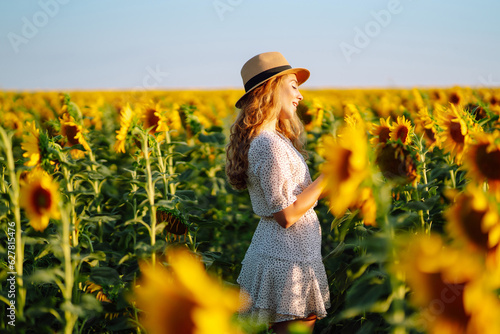 Sunny beautiful photo of young happy woman  enjoying warm weather  walking in blooming sunflower field. Woman poses in sunflower field in a dress and hat. Enjoy the moment. Active lifestyle.