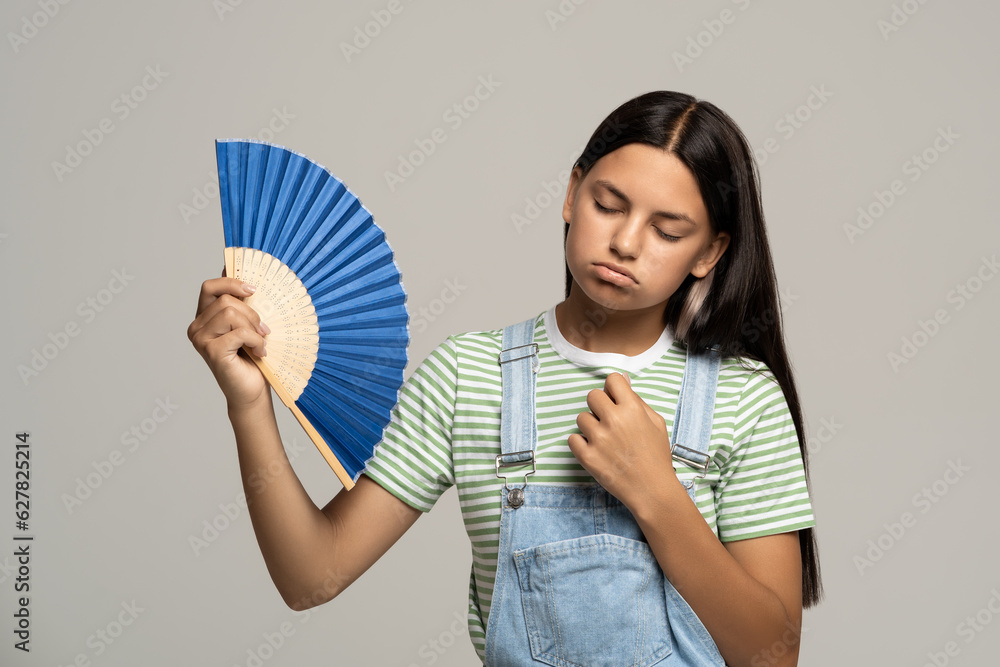 Sweaty overheated teenage girl using paper fan suffer from heat feels sluggish. Displeased tired teen girl in overalls cooling in hot summer weather isolated on studio gray background. Stuffiness. Sweaty overheated teenage girl using paper fan suffer from heat feels sluggish. Displeased tired teen girl in overalls cooling in hot summer weather isolated on studio gray background. Stuffiness.