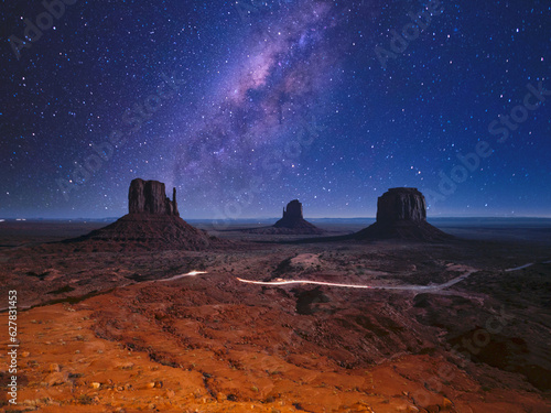 View of the 3 monoliths of Monument Valley at night, with starry sky and milky way, and a car passing on the road below