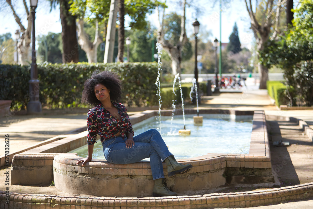 Young woman, beautiful and black with afro hair, with flower shirt, jeans and boots, sitting in a fountain, relaxed and quiet. Concept beauty, relaxation, tranquility, water.