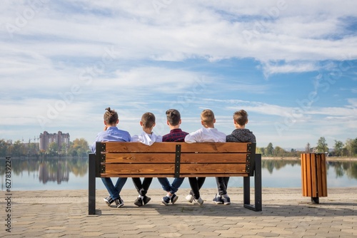 Five young boys are sitting on a wooden weasel near a pond. People rest on the bank of a river or lake. Youthful dreams or plans for the future. Teenagers have their backs turned to the camera. 