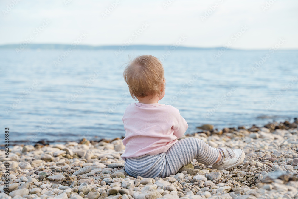 A one-year-old toddler sits on the beach of the sea in the evening after sunset A one-year-old toddler sits on the beach of the sea in the evening after sunset