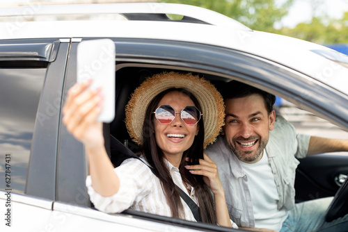 Happy young couple 30s taking selfie inside car, using phone