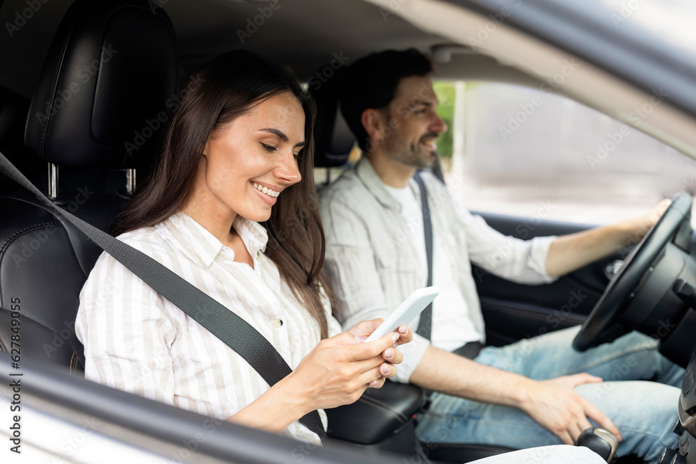 Happy millennial couple sitting inside white car, woman holding smartphone Happy millennial couple sitting inside white car, woman holding smartphone