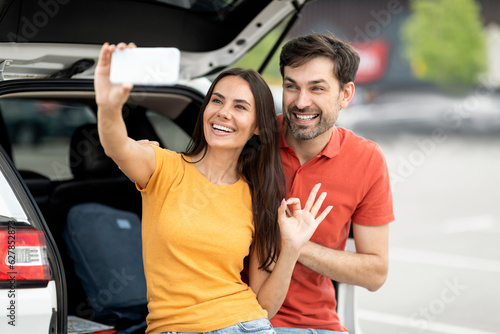 Happy couple travellers taking selfie next to open car trunk