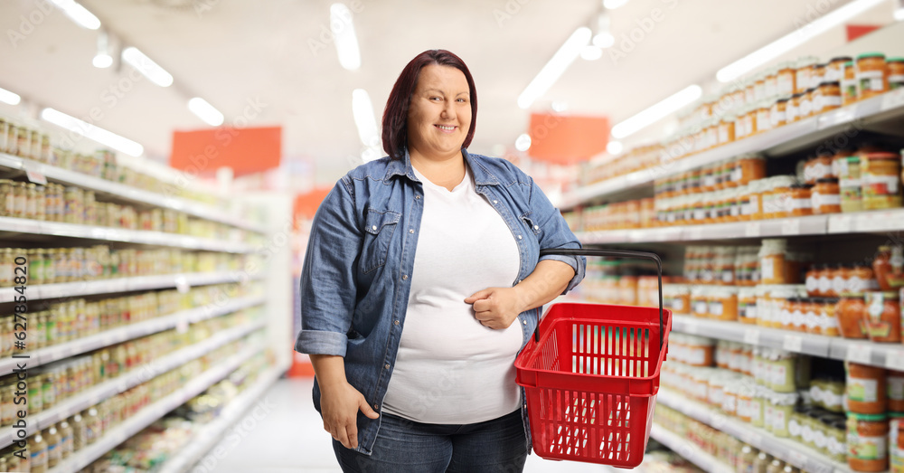 Corpulent woman with a shopping basket smiling and posing inside a supermarket