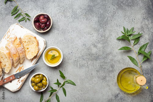 Sliced fresh ciabatta on cutting kitchen board, green and brown olives, olive oil with rosemary, olive tree branches on gray concrete stone rustic background from above copy space