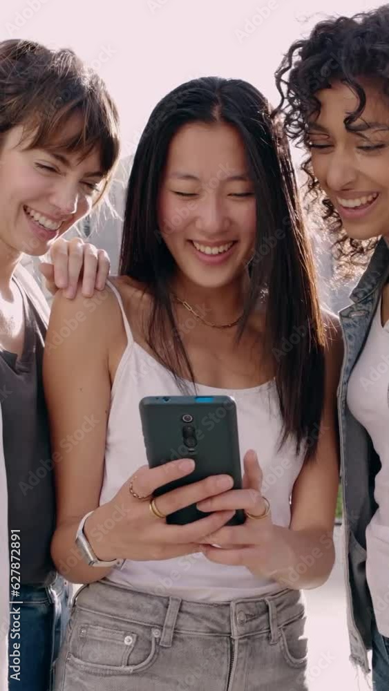 Three multiethnic young women having fun using social media app on mobile phone. Small group of female friends smile while looking at smartphone screen over city background. Vertical video. Three multiethnic young women having fun using social media app on mobile phone. Small group of female friends smile while looking at smartphone screen over city background. Vertical video.