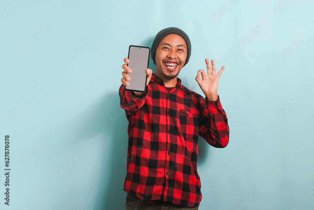 Young Asian man with a beanie hat and a red plaid flannel shirt shows an empty white blank display on his phone and makes the OK gesture, while standing against a blue background Young Asian man with a beanie hat and a red plaid flannel shirt shows an empty white blank display on his phone and makes the OK gesture, while standing against a blue background