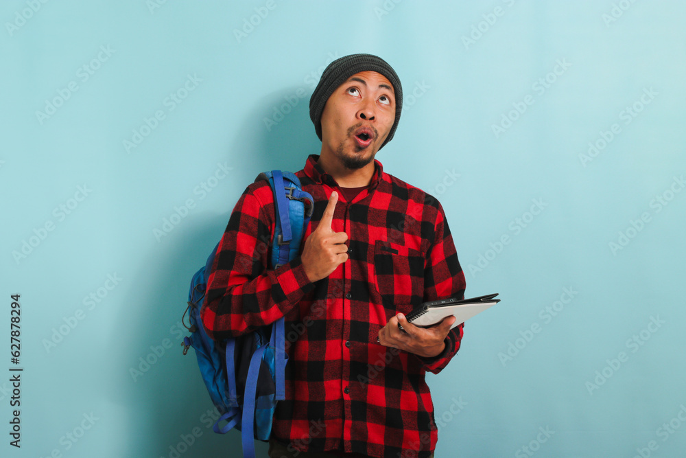 An Excited smiling young Asian student with a beanie hat and a red plaid flannel shirt, wearing a backpack, is pointing finger up at copy space while standing against a blue background An Excited smiling young Asian student with a beanie hat and a red plaid flannel shirt, wearing a backpack, is pointing finger up at copy space while standing against a blue background