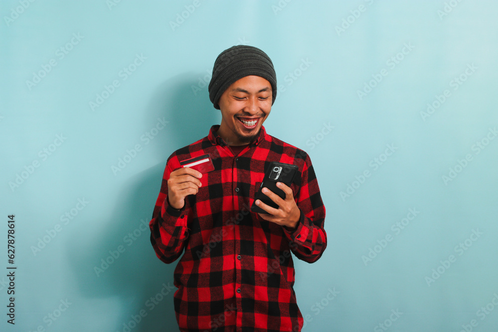 Happy young Asian man with a beanie hat and a red plaid flannel shirt is holding a smartphone and a banking credit card, doing online shopping, isolated on a blue background. Happy young Asian man with a beanie hat and a red plaid flannel shirt is holding a smartphone and a banking credit card, doing online shopping, isolated on a blue background.