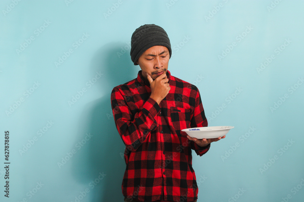 Pensive Young Asian man with beanie hat and red plaid flannel shirt is feeling confused, looking aside, thinking about what to eat while holding an empty white plate, isolated on a blue background Pensive Young Asian man with beanie hat and red plaid flannel shirt is feeling confused, looking aside, thinking about what to eat while holding an empty white plate, isolated on a blue background
