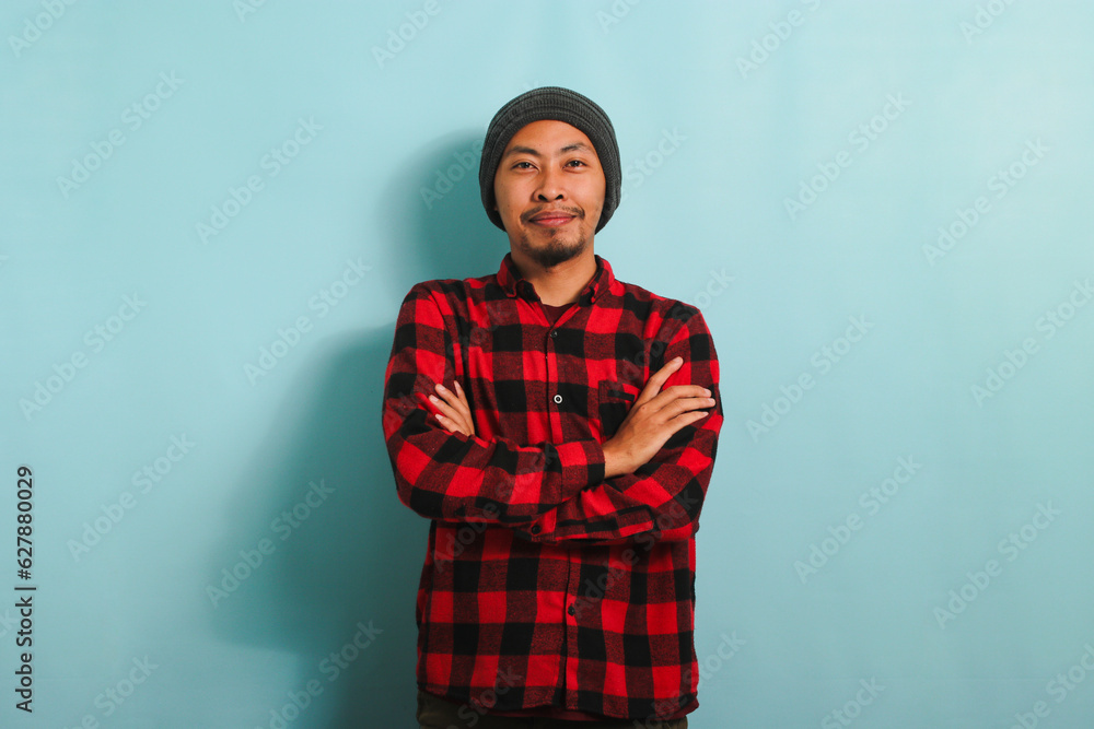 Fototapeta premium A confident young Asian man with a beanie hat and a red plaid flannel shirt is looking at the camera with his arms crossed. He is isolated on a blue background