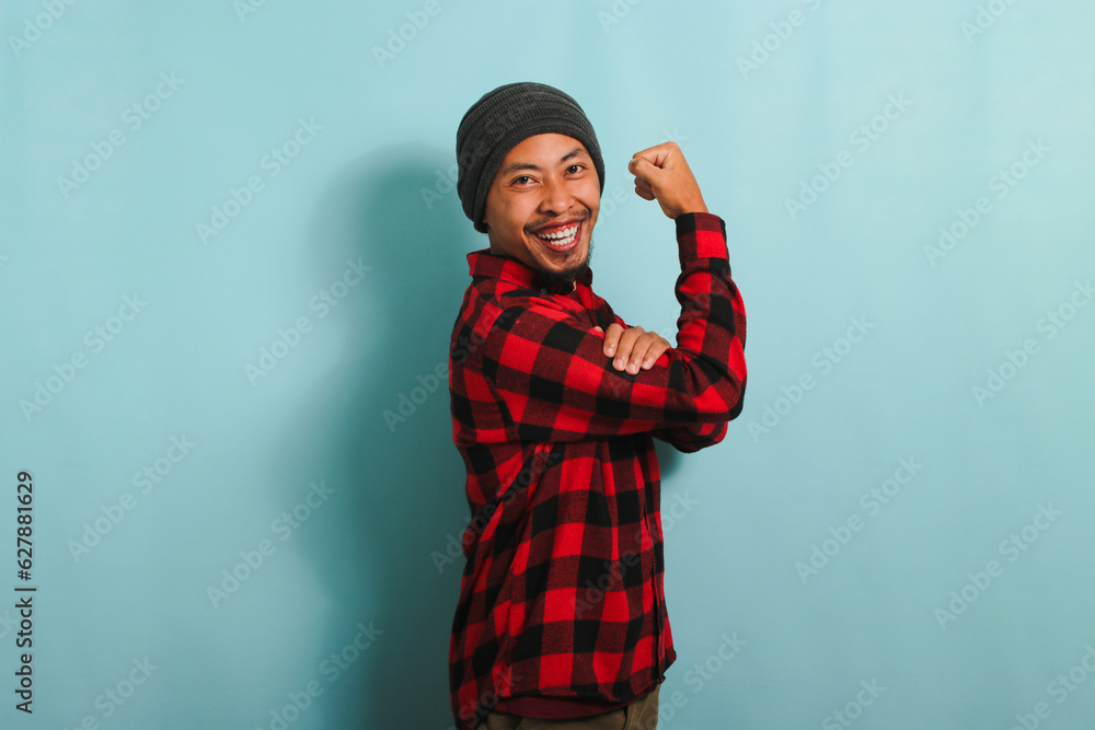 Excited Young Asian man with beanie hat and red plaid flannel shirt making a strong gesture, showing strength by flexing his arms and muscles, isolated on a blue background Excited Young Asian man with beanie hat and red plaid flannel shirt making a strong gesture, showing strength by flexing his arms and muscles, isolated on a blue background