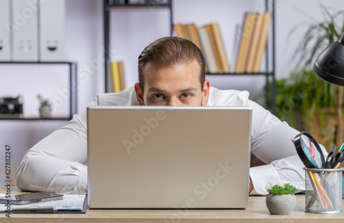 Lebanese confident businessman hiding behind laptop computer, looking at camera, spying his colleagues working, peeping. Professional freelancer man looking from behind computer with cunning eyes photo