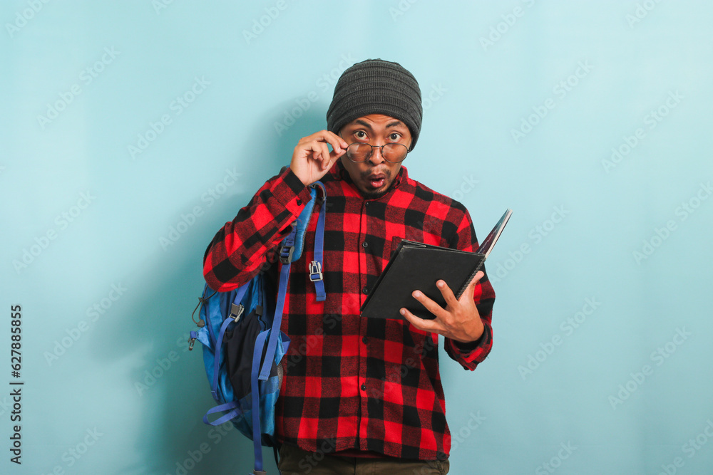 A surprised young Asian student in a beanie hat and a red plaid flannel shirt, wearing a backpack and glasses, is reading a book with an astonished expression while standing against a blue background A surprised young Asian student in a beanie hat and a red plaid flannel shirt, wearing a backpack and glasses, is reading a book with an astonished expression while standing against a blue background