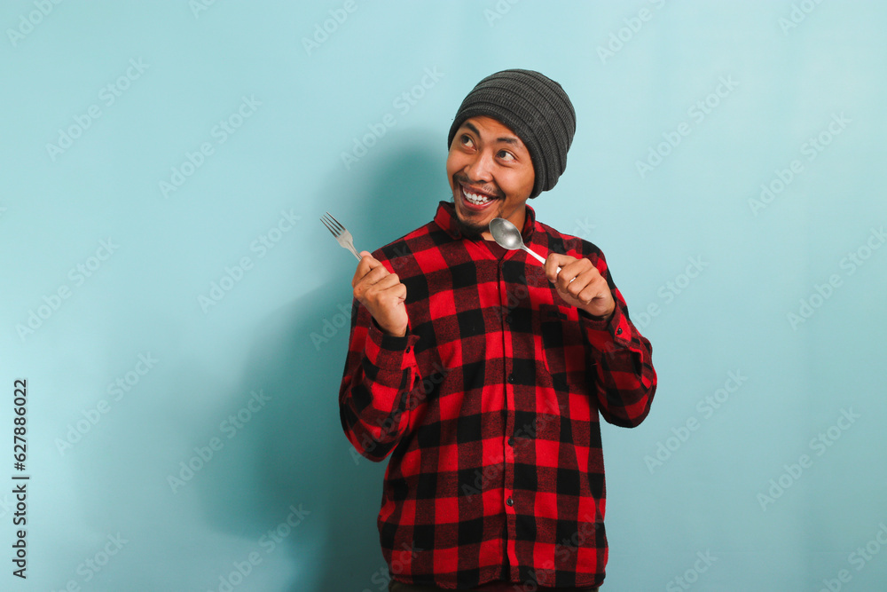 Excited Young Asian man with beanie hat and red plaid flannel shirt pointing aside at copy space with a spoon and fork, as if indicating a tempting meal or food option, isolated on a blue background Excited Young Asian man with beanie hat and red plaid flannel shirt pointing aside at copy space with a spoon and fork, as if indicating a tempting meal or food option, isolated on a blue background