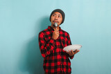 Pensive Young Asian man with beanie hat and red plaid flannel shirt holding a spoon and an empty white plate in his hand, thinking about what to eat, isolated on a blue background