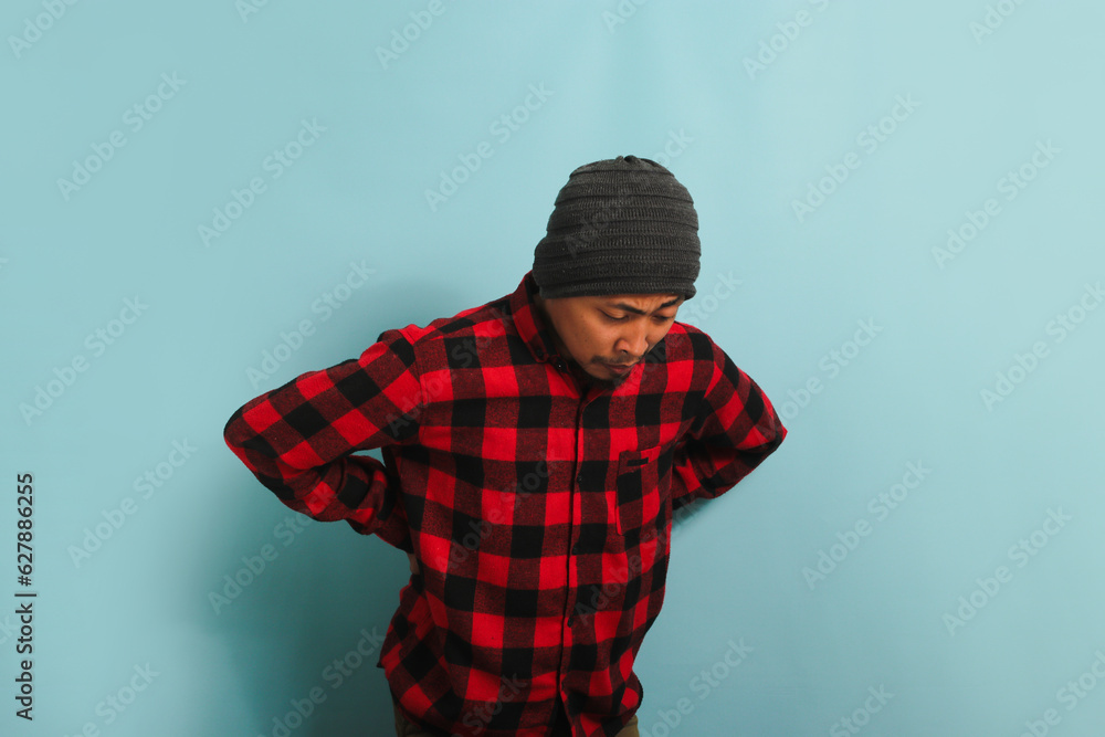 A young Asian man with a beanie hat and a red plaid flannel shirt is suffering from a backache, experiencing back pain. He is isolated on a blue background