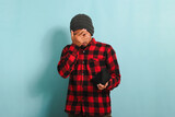 Funny young Asian man with beanie hat and red plaid flannel shirt covering his face with his hand and peeking between his fingers while looking at his empty wallet, isolated on a blue background