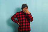 A young Asian man wearing a beanie hat and a red plaid flannel shirt is sneezing into a tissue, suffering from a cough, fever, and flu. He is isolated on a blue background