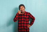 An excited young Asian man wearing a beanie hat and a red plaid flannel shirt makes a CALL ME gesture with his hand, isolated on a blue background