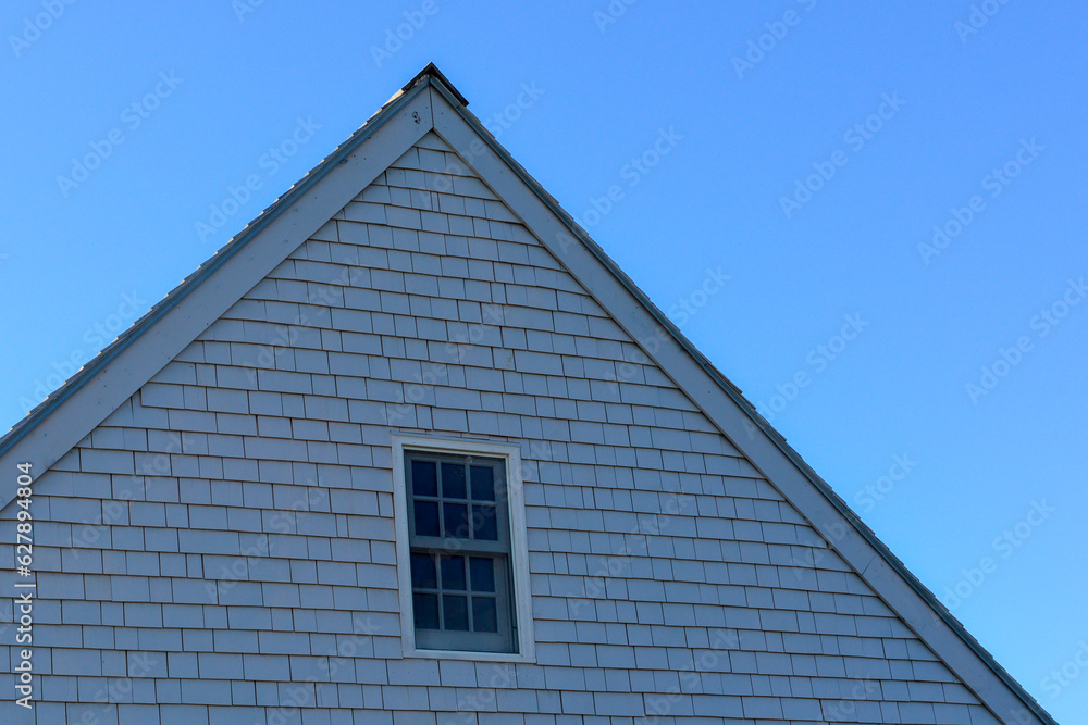 The exterior wall of a worn grey and weathered cedar shingled house with a small closed double hung window with blue and white trim. The tall vintage building has a clear blue sky in the background.