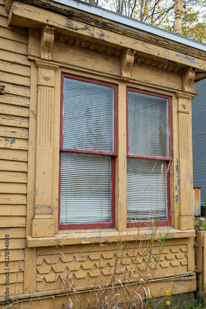 The exterior wall of a yellow wooden house with a large double hung window. The glass window has a curtain and blind. The pattern on the outside of the wall is small scallop shapes and horizontal wood
