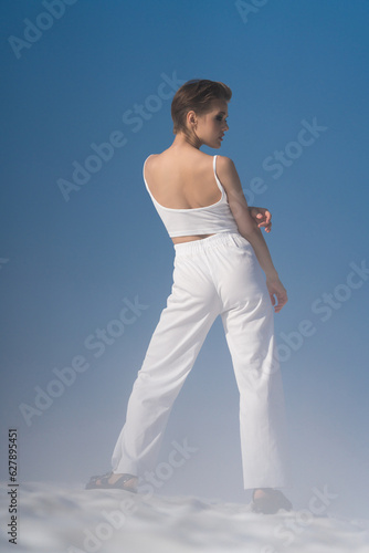 Full length back view of sensual fashion model standing through snow with light fog on sunny weather with blue sky. Sexual blond woman with short hair dressed in white crop top, white pants, sandals