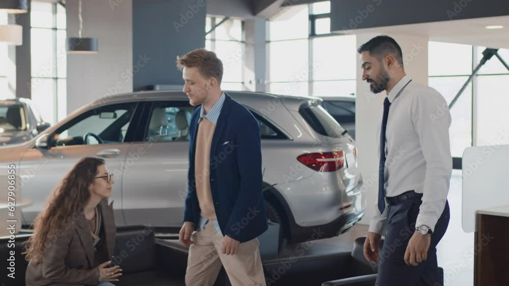 Full shot of polite Middle Eastern car salesman welcoming young Caucasian couple into showroom, inviting to sit on couch, shaking hands with husband, and giving them copies of loan agreement to read Full shot of polite Middle Eastern car salesman welcoming young Caucasian couple into showroom, inviting to sit on couch, shaking hands with husband, and giving them copies of loan agreement to read