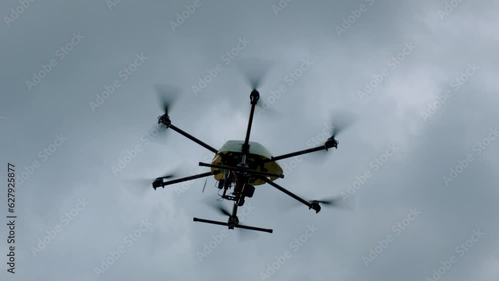 From below, handheld camera captures black and yellow drone with a camera, six propellers, soaring against a cloudy sky From below, handheld camera captures black and yellow drone with a camera, six propellers, soaring against a cloudy sky