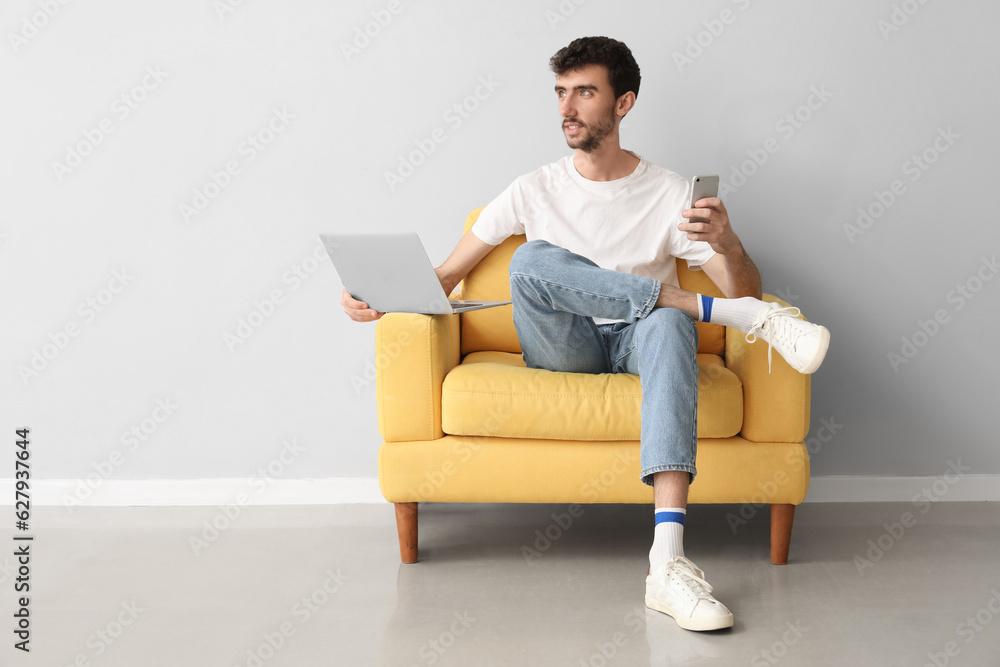 Young man with laptop and mobile phone in armchair near light wall Young man with laptop and mobile phone in armchair near light wall