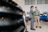 Remote low-angle view of male client talking to car dealer in business suit in dealership discussing automobiles looking at luxurious new model. Concept of choosing and buying new auto at showroom.