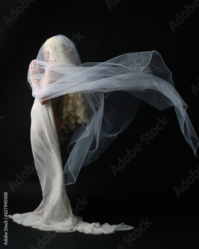 Full length portrait of beautiful blonde woman wearing white gown dress with flowing ghostly veiled fabric, isolated on dar studio background.