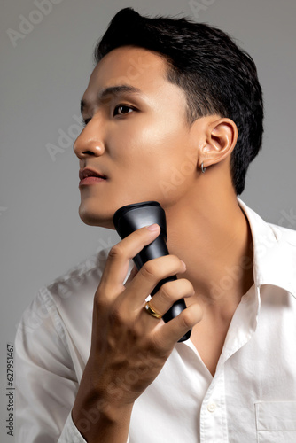 Studio portrait of young handsome Asian man with nice facial skin shaving with electric razor machine on light gray isolated background. photo