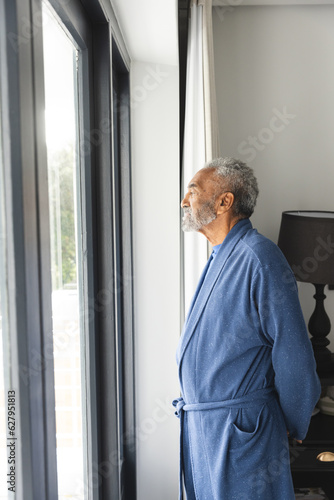 Thoughtful senior biracial man wearing bathrobe and looking through window at home