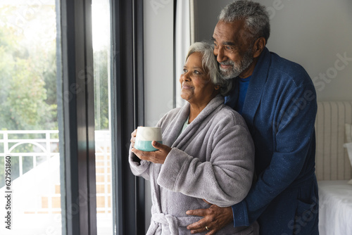 Happy senior biracial couple wearing bathrobes and looking through window with mug of coffee at home