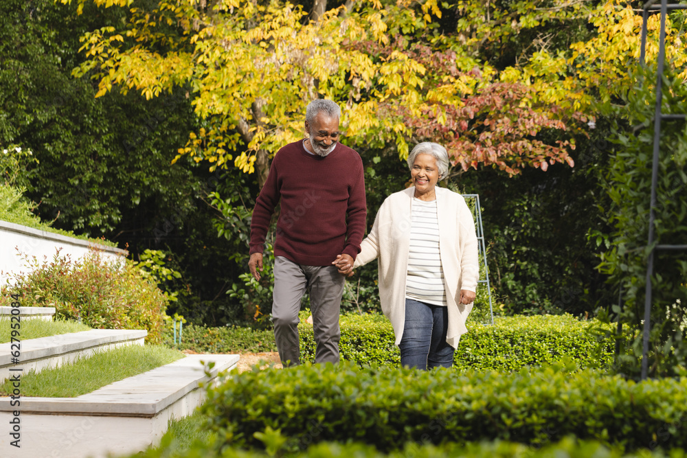 Obraz premium Happy senior biracial couple holding hands and walking in garden at home